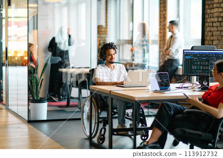 A young business group, including an African American businessman in a wheelchair, collaborates within a modern glass office, actively engaged around a computer and laptop, collectively solving A young business group, including an African American businessman in a wheelchair, collaborates within a modern glass office, actively engaged around a computer and laptop, collectively solving 113912732