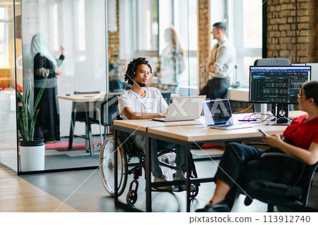 A young business group, including an African American businessman in a wheelchair, collaborates within a modern glass office, actively engaged around a computer and laptop, collectively solving 113912740