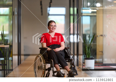 A modern young businesswoman in a wheelchair is surrounded by an inclusive workspace with glass-walled offices, embodying determination and innovation in the business world 113912801