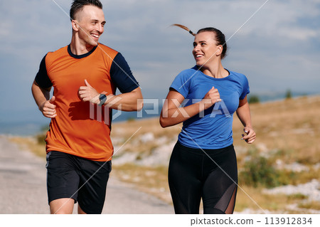 A couple dressed in sportswear runs along a scenic road during an early morning workout, enjoying the fresh air and maintaining a healthy lifestyle A couple dressed in sportswear runs along a scenic road during an early morning workout, enjoying the fresh air and maintaining a healthy lifestyle 113912834