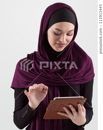 Beautiful and modern young Arabic businesswoman in hijab using a tablet computer while posing on light background and smiling at the camera. Business diversity concept, Muslim lady. 113913443