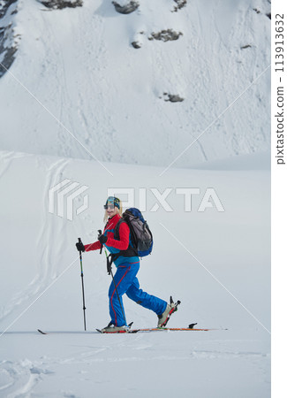 A female skier stands at the snowy summit of a mountain, equipped with professional gear and skis, poised for an exhilarating descent. 113913632