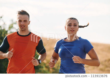 A couple dressed in sportswear runs along a scenic road during an early morning workout, enjoying the fresh air and maintaining a healthy lifestyle A couple dressed in sportswear runs along a scenic road during an early morning workout, enjoying the fresh air and maintaining a healthy lifestyle 113913676