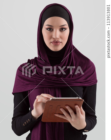 Beautiful and modern young Arabic businesswoman in hijab using a tablet computer while posing on light background and smiling at the camera. Business diversity concept, Muslim lady. Beautiful and modern young Arabic businesswoman in hijab using a tablet computer while posing on light background and smiling at the camera. Business diversity concept, Muslim lady. 113913801