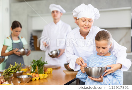 During childrens cooking class, female chef helps girl thoroughly stir homogeneous dough with whisk 113917891