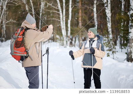 Medium long shot of senior Caucasian tourist holding smartphone taking photo of his wife in forest park on winter day 113918853