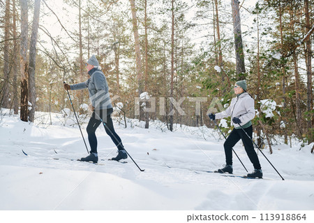 Side view wide shot of senior man and woman in sportswear spending time together skiing in mountain forest on winter day, copy space 113918864
