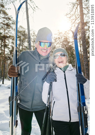 Vertical medium long shot of joyful active senior couple in winter sportswear and sunglasses standing outdoors posing for camera with skis and poles 113918895