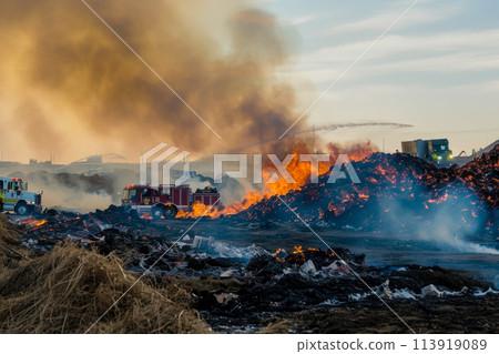 landfill burning. The fire is out of control, and there are firefighters and trucks in the background 113919089