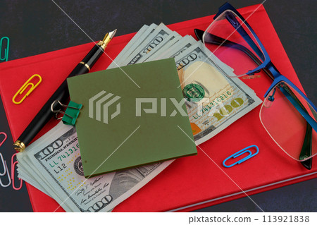 A sticker for writing attached to dollars on a red business notebook, glasses next to it, a pen on a dark background 113921838