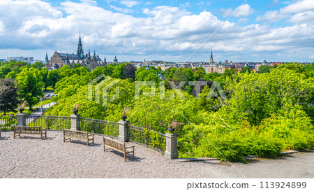 A peaceful view from King Oscar Terrace in Skansen showing wooden benches, lush trees, and distant city architecture under a bright blue sky. Stockholm, Sweden A peaceful view from King Oscar Terrace in Skansen showing wooden benches, lush trees, and distant city architecture under a bright blue sky. Stockholm, Sweden 113924899