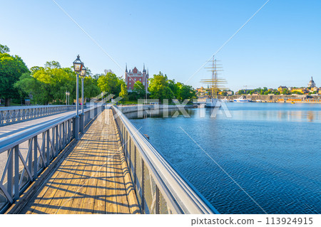 A vibrant day at Skeppsholmen Bridge in Stockholm, with clear skies and sunlight bathing the walkway, the calm water, and historic buildings in the distance. Stockholm, Sweden 113924915