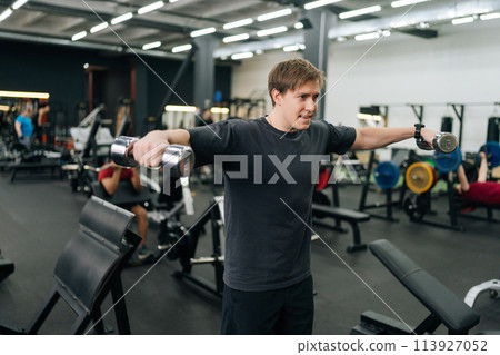 Portrait of motivated beginner sportsman hard time doing lateral arm raise exercise with dumbbells. Tired male athlete improves shoulders with dumbbell breeding. Concept of healthy lifestyle 113927052