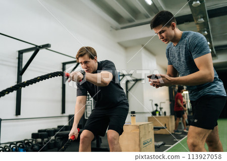 Portrait of dedicated sportsman exercising with battle rope while coach encouraging during cross training in health club standing with stopwatch. Concept of healthy lifestyle, physical sport activity. Portrait of dedicated sportsman exercising with battle rope while coach encouraging during cross training in health club standing with stopwatch. Concept of healthy lifestyle, physical sport activity. 113927058