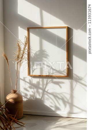 A white framed picture sits on a wall next to a vase of dried grass 113931674