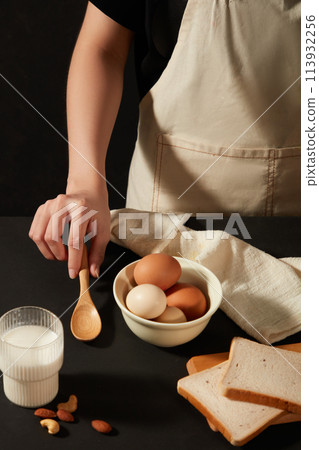 Beauty hand is putting a wooden spoon on table, some eggs are contained on a round bowl with a cream towel, some sandwiches besides, a cup of fresh nut milk. Cooking concept for culinary products 113932256