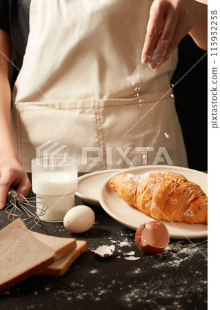 Flour is sprinkled into a cake contained on a round dish by male hand next to a glass of milk, egg and some sandwiches on dark color kitchen table. Blank space for designing Flour is sprinkled into a cake contained on a round dish by male hand next to a glass of milk, egg and some sandwiches on dark color kitchen table. Blank space for designing 113932258