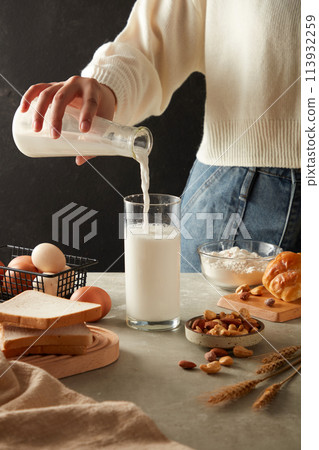 Female hand is pouring fresh milk into a glass decorated with some sandwiches, eggs, nuts, a bowl of flour and a cake on kitchen background. Advertising photo for cooking concept Female hand is pouring fresh milk into a glass decorated with some sandwiches, eggs, nuts, a bowl of flour and a cake on kitchen background. Advertising photo for cooking concept 113932259
