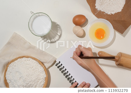 Culinary concept photo with a female hand holding a pencil to write recipe on a notebook, surrounded by some baking pastry ingredients. Space for design or displaying elements Culinary concept photo with a female hand holding a pencil to write recipe on a notebook, surrounded by some baking pastry ingredients. Space for design or displaying elements 113932270