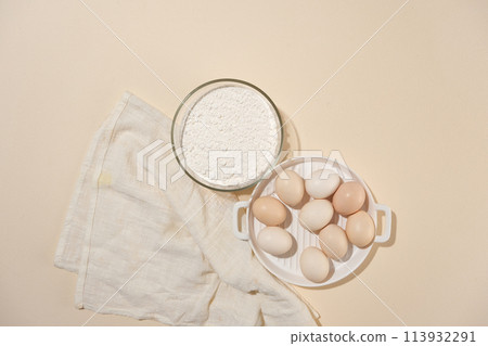Top view with a glass bowl off flour and plenty of eggs lay flat on pink table, decorated with a white towel. Cooking layout for advertising product made of egg and flour Top view with a glass bowl off flour and plenty of eggs lay flat on pink table, decorated with a white towel. Cooking layout for advertising product made of egg and flour 113932291