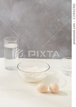Basic culinary concept with a bowl of flour, some eggs lay on the table and a glass of fresh water on gray marble background. Blank space for adding text and presenting product Basic culinary concept with a bowl of flour, some eggs lay on the table and a glass of fresh water on gray marble background. Blank space for adding text and presenting product 113932302