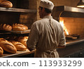 A baker inspects freshly baked bread in a bakery kitchen. The warm lighting emphasizes the quality and care in his work. 113933299