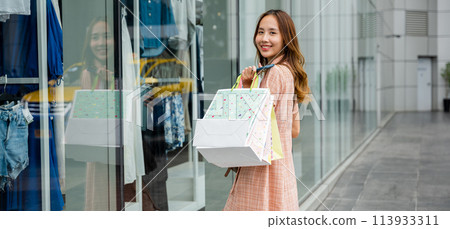 A young woman walks down the street, holding shopping bags and looking into a clothing store window display. Fashion trends are always in style A young woman walks down the street, holding shopping bags and looking into a clothing store window display. Fashion trends are always in style 113933311