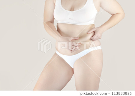 Woman has excess fat. Girl in underwear shows belly fat. She wants to lose weight nude beige background Woman has excess fat. Girl in underwear shows belly fat. She wants to lose weight nude beige background 113933985