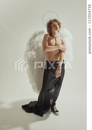 Young man with large wings and halo posing wrapped in black fabric looking away against white studio background. 113934736
