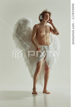 Young man with large wings looks and angel listening music in modern headphones against white studio background. 113934840