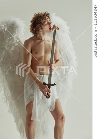Young man with curly hair and wings poses with sword from which he eats raw meat against white studio background. Young man with curly hair and wings poses with sword from which he eats raw meat against white studio background. 113934847
