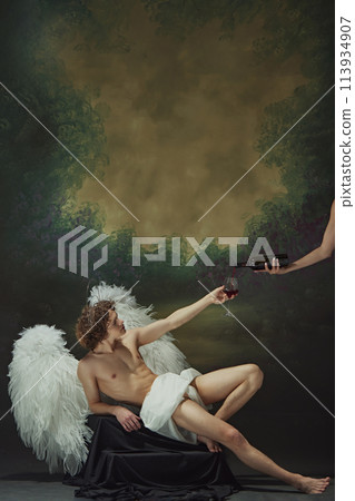 Man with curly hair sits with large wings spread out behind him, reaching out to hand to fill glass with wine against vintage studio background. Man with curly hair sits with large wings spread out behind him, reaching out to hand to fill glass with wine against vintage studio background. 113934907