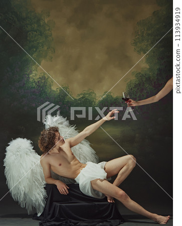 Man with curly hair sits with large wings spread out behind him, reaching out to hand with glass of wine against vintage studio background. 113934919