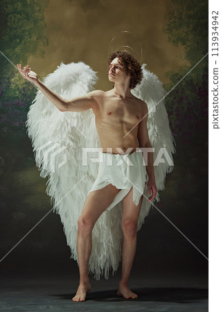 Celestial Elegance. Strikingly handsome man with curly hair and angelic wings poses effortlessly against vintage studio background. 113934942
