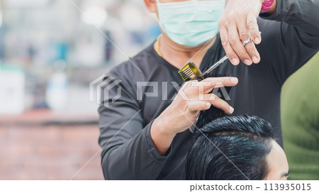 professional man hairdresser wearing face mask holding scissors and haircutting client woman indoor salon, man make business hair care has tool haircut to service haircut to customer, selective focus 113935015