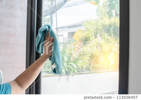 The woman is using a cloth and a glass cleaner to clean the glass door to remove the dirt. A female wearing a mask to prevent the coronavirus or COVID-19. Close up and selective focus on hands. The woman is using a cloth and a glass cleaner to clean the glass door to remove the dirt. A female wearing a mask to prevent the coronavirus or COVID-19. Close up and selective focus on hands. 113936407