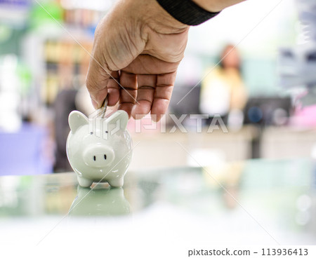 Close up. Hand of a man holding a coin for putting to white piggy bank in the bank office. Money-saving concept investment budget wealth business. Copy space on design, text. Bokeh, blurred background Close up. Hand of a man holding a coin for putting to white piggy bank in the bank office. Money-saving concept investment budget wealth business. Copy space on design, text. Bokeh, blurred background 113936413