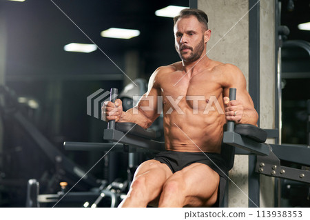 Handsome bearded sportsman pulling up, raising legs, working out indoors. Front view of muscular male athlete with bare torso doing pull ups on training apparatus in gym. Concept of sport, workout. 113938353