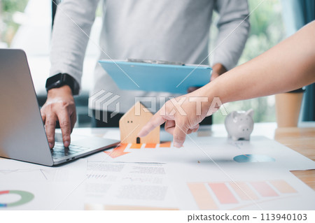 A man is pointing at a house on a table with a laptop and a blue folder 113940103