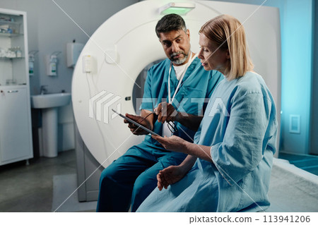Biracial radiographer sitting on CT scanner bed showing X-ray image to mature Caucasian female patient, copy space Biracial radiographer sitting on CT scanner bed showing X-ray image to mature Caucasian female patient, copy space 113941206