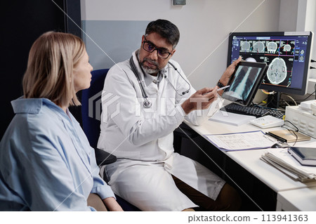 Serious biracial radiographer showing chest X-ray image to mature Caucasian female patient after MRI procedure, copy space 113941363