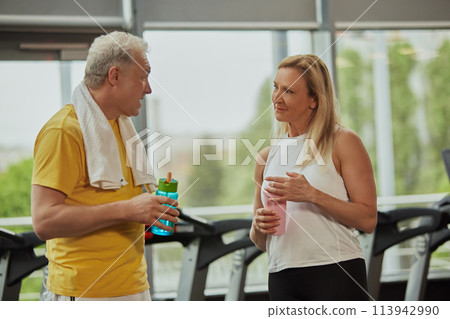 Senior man and woman in sportswear with water bottles taking break during gym time at fitness center. Senior man and woman in sportswear with water bottles taking break during gym time at fitness center. 113942990
