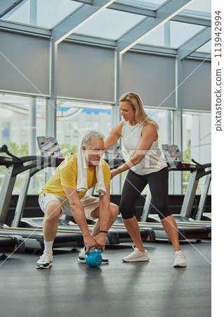 Senior man doing exercise with kettlebell and woman insures him in modern fitness center for retirements. Mature people in modern lifestyle. Senior man doing exercise with kettlebell and woman insures him in modern fitness center for retirements. Mature people in modern lifestyle. 113942994