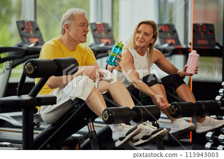 Two seniors, man and woman in sportswear share fun moment while resting between exercises in modern gym. 113943003