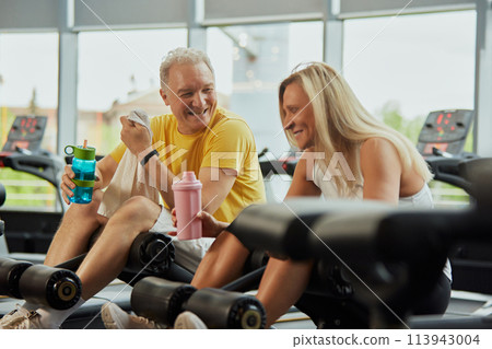 Two seniors in wearing sport gear share fun moment during exercising in modern fitness center. Active seniors in modern life Two seniors in wearing sport gear share fun moment during exercising in modern fitness center. Active seniors in modern life 113943004