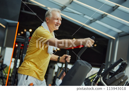 Positive, smiling mature man in vibrant sports attire with towel on shoulders work out on exercise machine at modern gym. 113943018