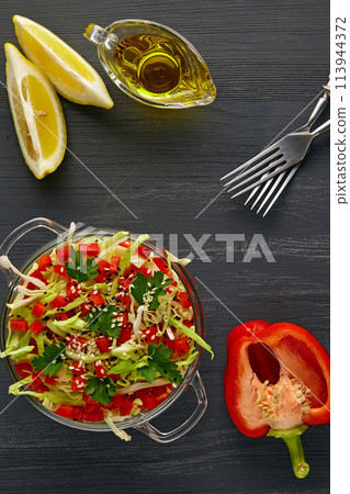 Vegetable salad of raw cabbage and bell pepper, dressed with olive oil and lemon, surrounded by ingredients on a black concrete table 113944372