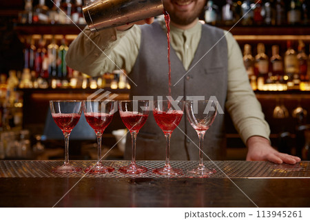 Bartender pouring freshly prepared cocktail into several glasses 113945261