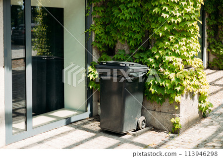A black trash can sitting next to a wall. Ecology concept. Help to reduce waste, Help global warming. Garbage bin A black trash can sitting next to a wall. Ecology concept. Help to reduce waste, Help global warming. Garbage bin 113946298