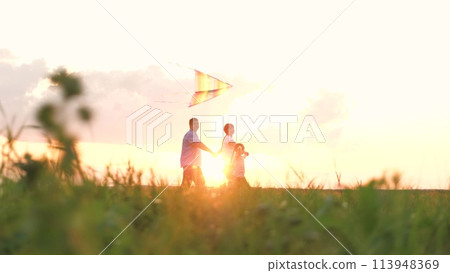 Cute little girl with flying kite mother father holding hands walking at sunset field together. Happy family parents and daughter outdoor playing flight toy entertainment having fun at nature meadow Cute little girl with flying kite mother father holding hands walking at sunset field together. Happy family parents and daughter outdoor playing flight toy entertainment having fun at nature meadow 113948369